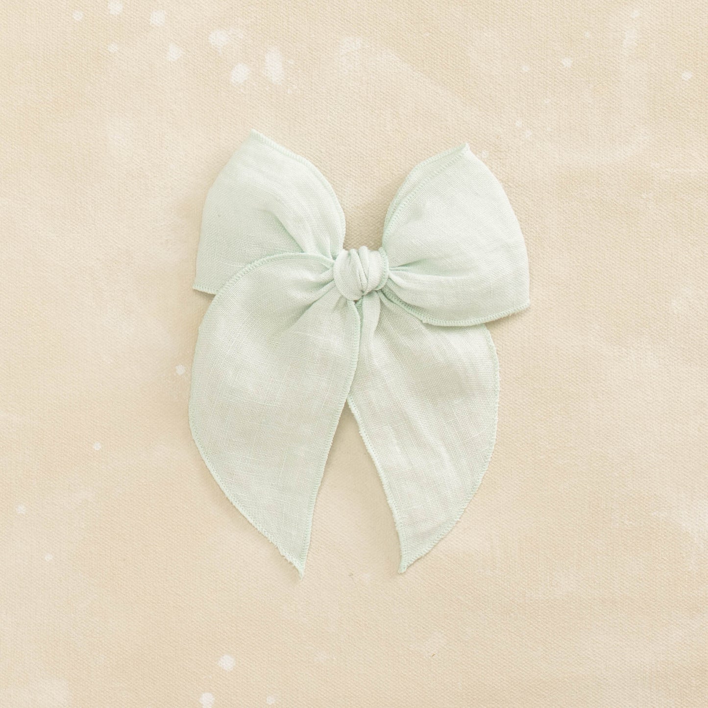 The Fleur // Tidewater by Shop Acre Goods is a light mint green fabric bow with a knotted center, smooth, symmetrical loops and tails, secured by an alligator clip against a cream-colored, textured background.