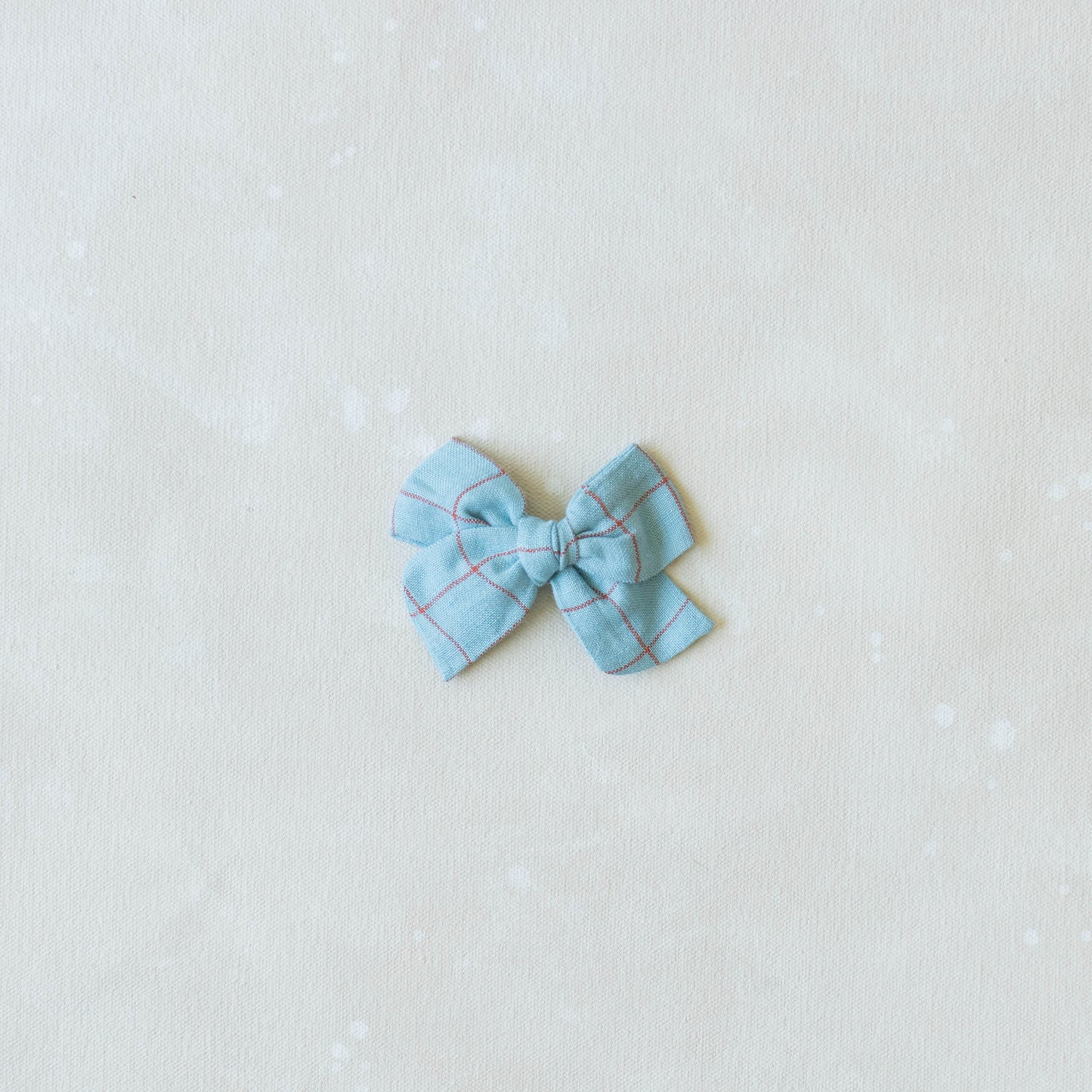 A small, light blue Explorer // Hopscotch bow by Shop Acre Goods features a red line grid pattern. Its mounted on a nylon headband and centered on a plain, light-colored background.