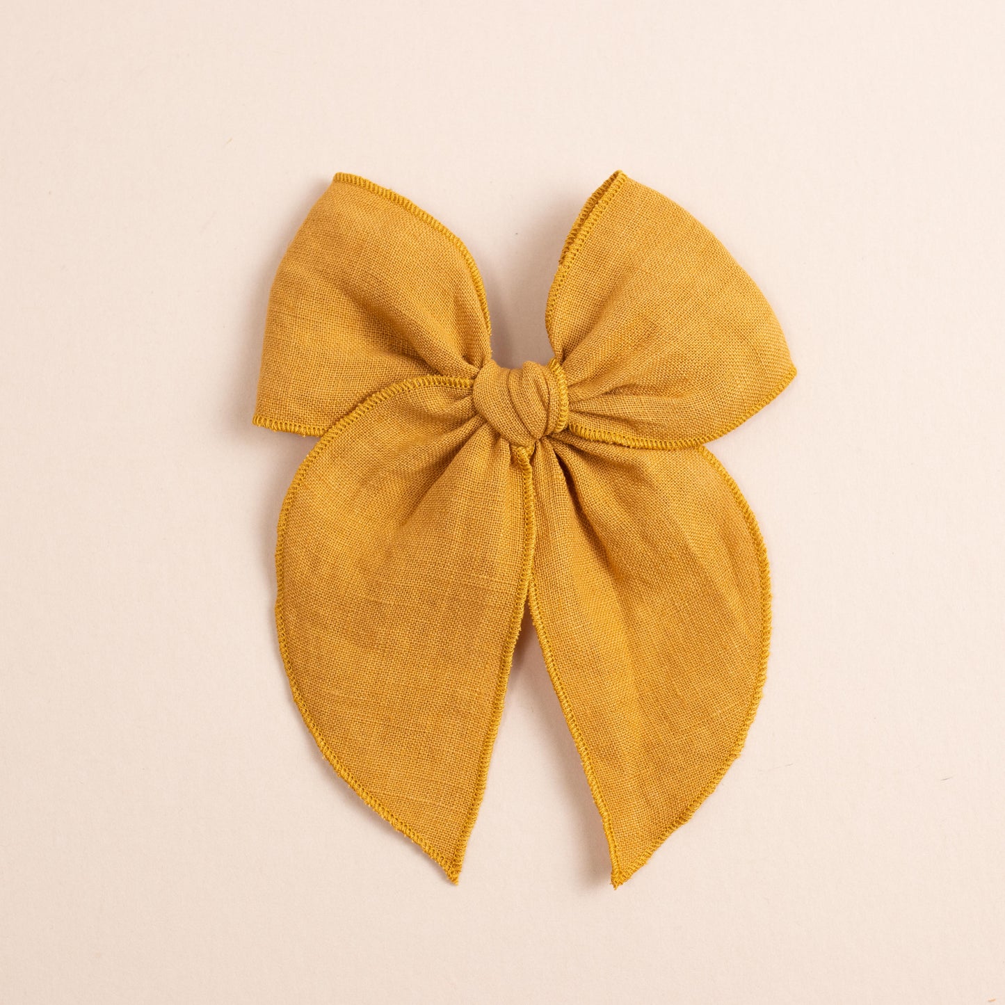 Placed against a soft beige background is the Fleur // No. 2 by Shop Acre Goods—a large, textured mustard yellow fabric bow with wide loops, long tails, and a neat hemmed edge.
