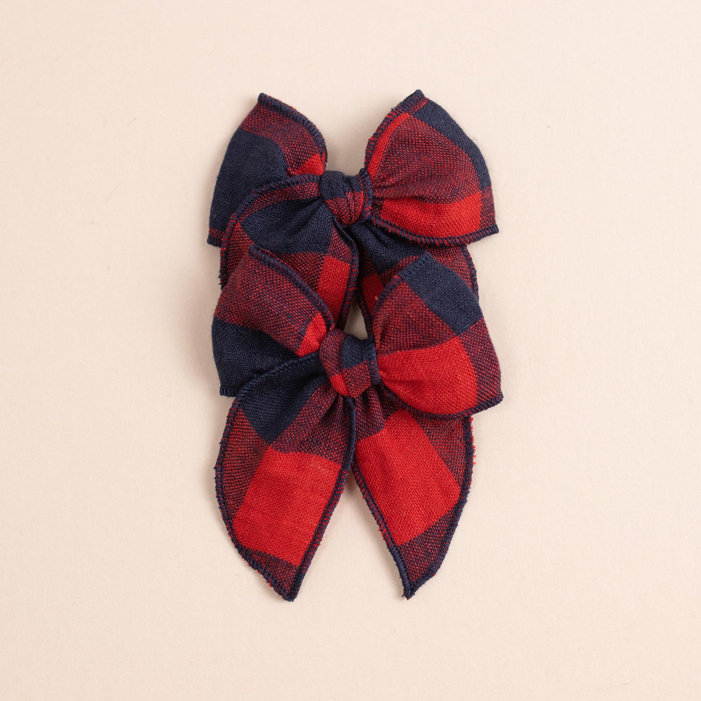 Two Mini Fleur // Game Day bows from Shop Acre Goods are arranged vertically on a light beige background. These red and navy plaid bows have knot centers, pointed tails, and attach with alligator clips for a rustic look. Ideal for adding charm to any ensemble.
