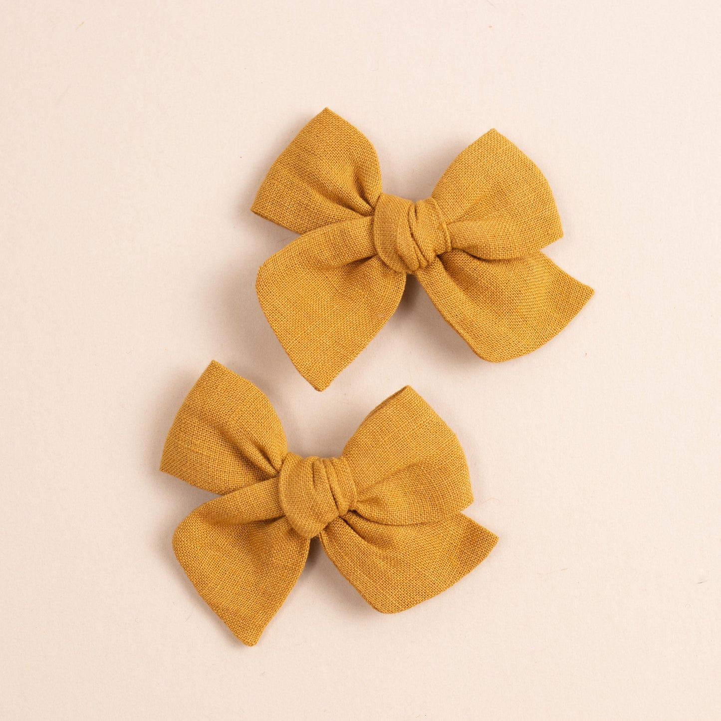 Two mustard-colored Explorer // No. 2 fabric bow ties by Shop Acre Goods rest on a plain beige background, ideal for a piggy set. Each bow features a central knot with symmetrical loops and tails, with the woven texture adding charm to any ensemble.