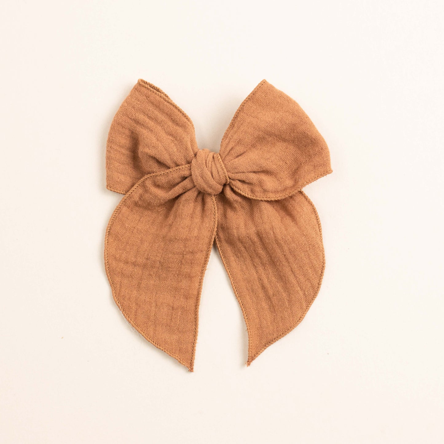 The Fleur // Tawny by Shop Acre Goods is a beige fabric bow with a textured finish against an off-white background, knotted at the center with hemmed edges and two long tails hanging symmetrically.