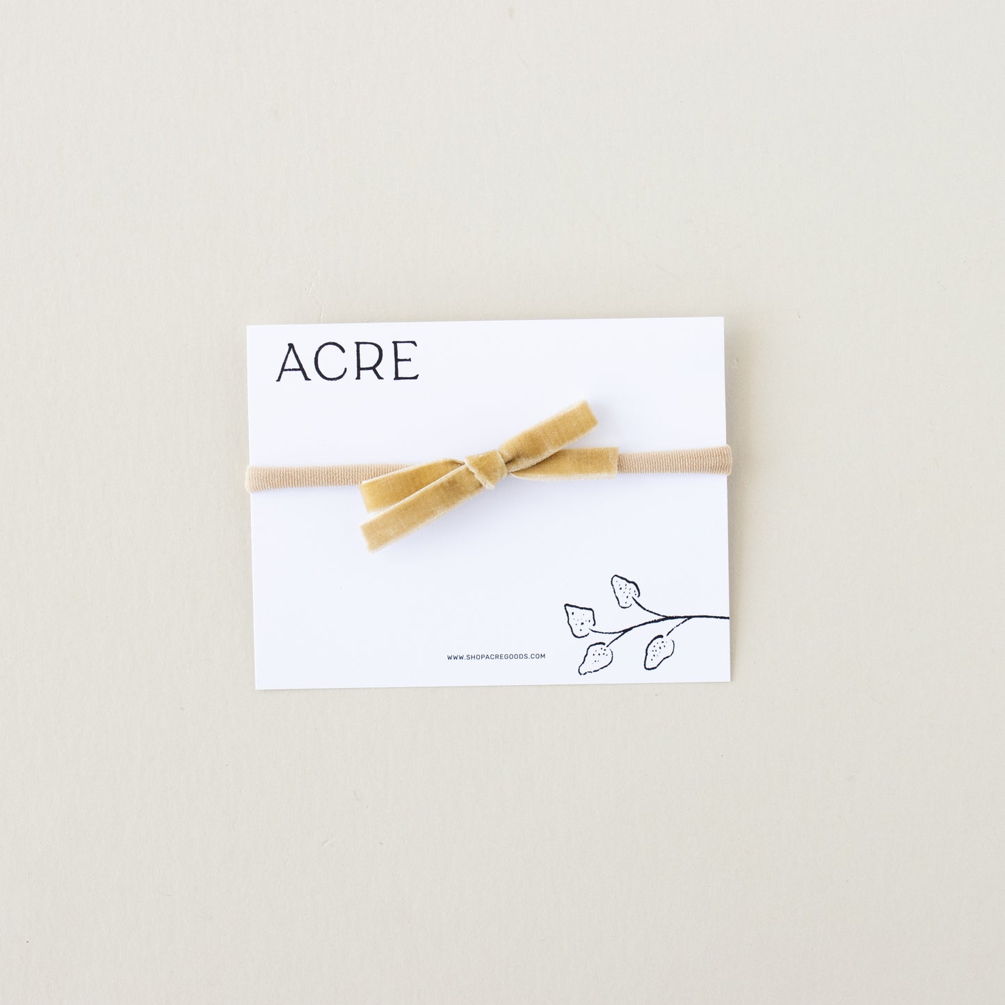 Displayed on a cream background, the hair accessory is a velvet ribbon bow in Pale Gold on a white card. ACRE and leaf illustrations adorn the card, representing Shop Acre Goods Velvet Infant Bow.