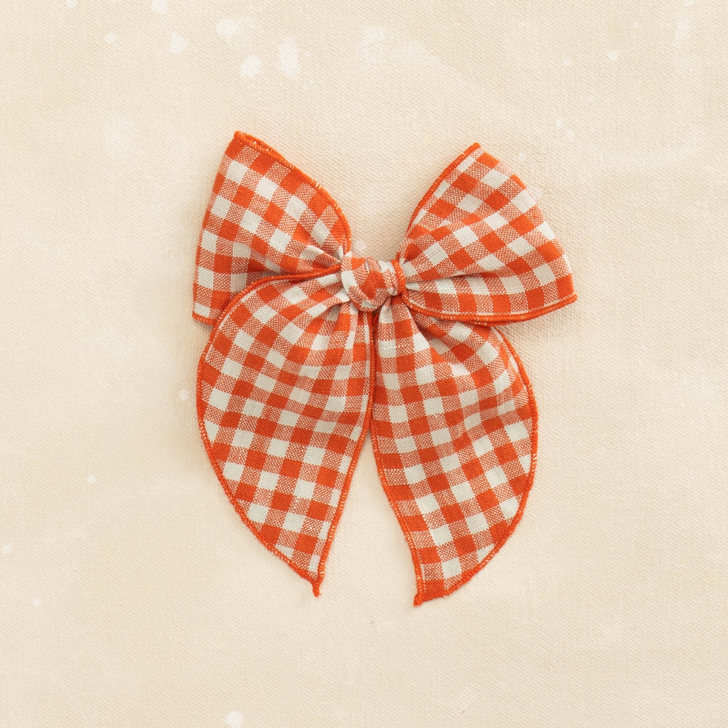 The Fleur // Whiplash by Shop Acre Goods features a tomato red and white gingham linen bow with long tails on a textured beige background.