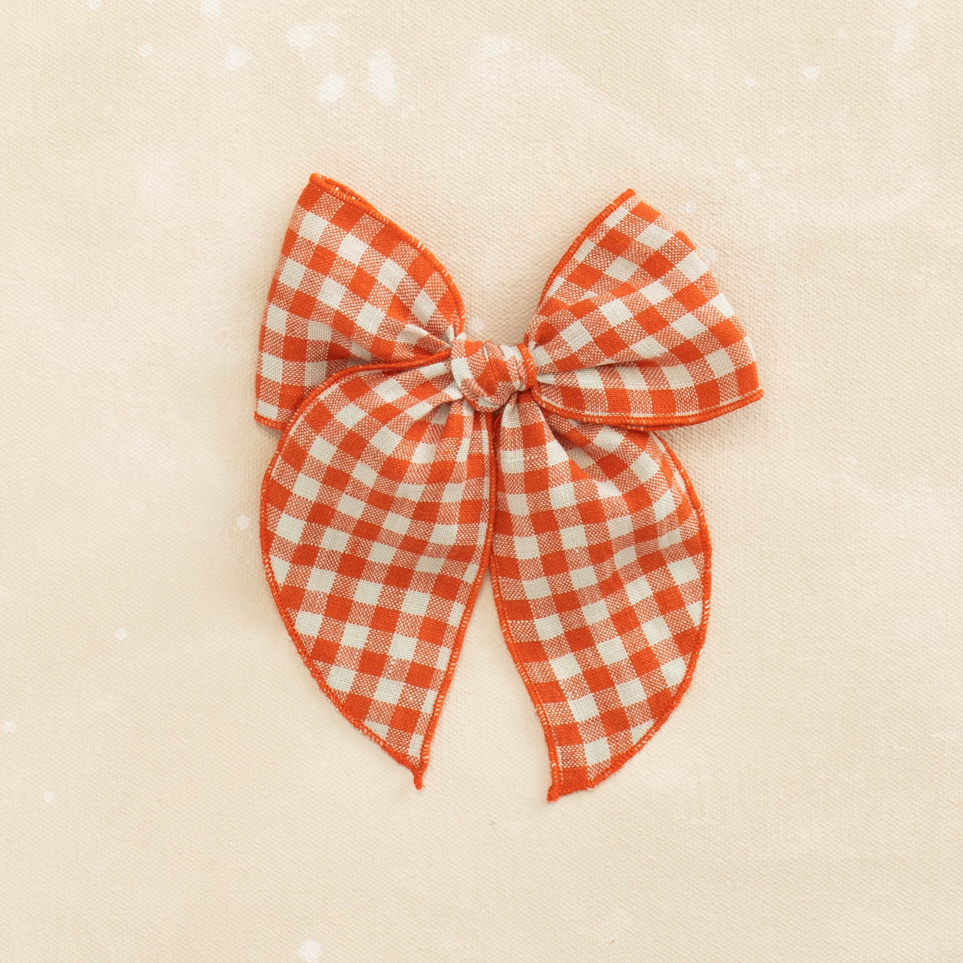 The Fleur // Whiplash by Shop Acre Goods features a tomato red and white gingham linen bow with long tails on a textured beige background.