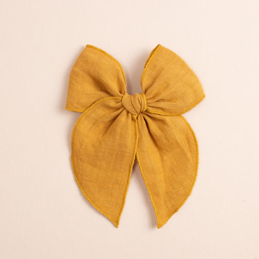 Placed against a soft beige background is the Fleur // No. 2 by Shop Acre Goods—a large, textured mustard yellow fabric bow with wide loops, long tails, and a neat hemmed edge.