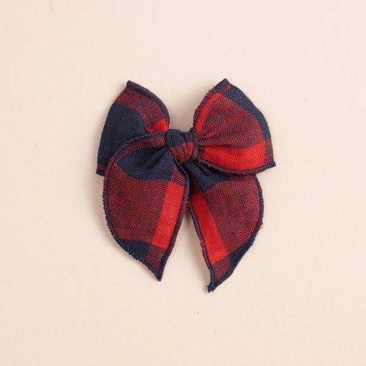 The Mini Fleur // Game Day by Shop Acre Goods is a large plaid bow with a red and blue checkered pattern, symmetrically designed on a plain beige background, seamlessly attached to a versatile nylon headband.