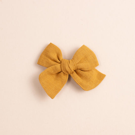 The Explorer // No. 2 by Shop Acre Goods is a mustard yellow fabric bow with a textured look, neatly tied to an alligator clip, and set against a light cream background.